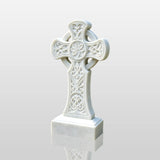 A white marble Celtic cross headstone rests on gravel among green plants.