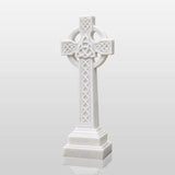 White marble Celtic cross tombstone stands on gravel ground surrounded by green plants.