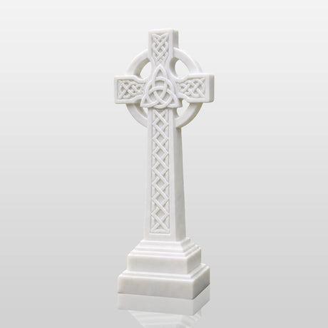 White marble Celtic cross tombstone stands on gravel ground surrounded by green plants.