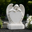 White marble tombstone carved with angel, roses and central blank heart stands on the black gravel cemetery ground.