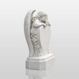 White marble tombstone carved with angel, roses and central blank heart stands on the black gravel cemetery ground.