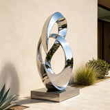 A stainless steel intertwined loop sculpture stands beside a walkway in front of a building.