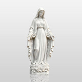 White marble our lady of Grace statue stands on a stone pathway near a church yard.