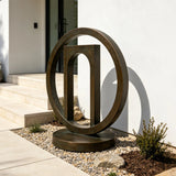 The geometric bronze sculpture stands in gravel planting bed near a home entrance.