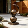 A bronze sculpture with spheres and a ring stands on concrete patio outside a modern house.