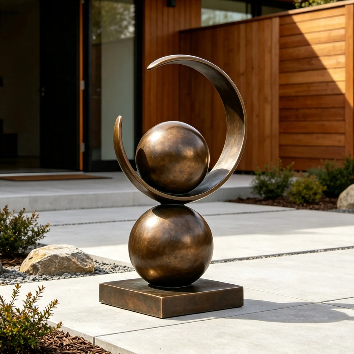 A bronze sculpture with spheres and a ring stands on concrete patio outside a modern house.