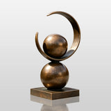 A bronze sculpture with spheres and a ring stands on concrete patio outside a modern house.