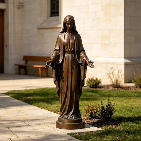 The bronze our lady of grace statue stands outdoors beside stone walkway near a church.