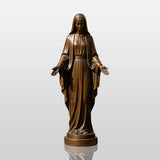 The bronze our lady of grace statue stands outdoors beside stone walkway near a church.