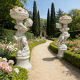 Pair of marble sculptures depicting children holding flowerpots line a curved gravel garden path beside trimmed hedges and rose blooms.