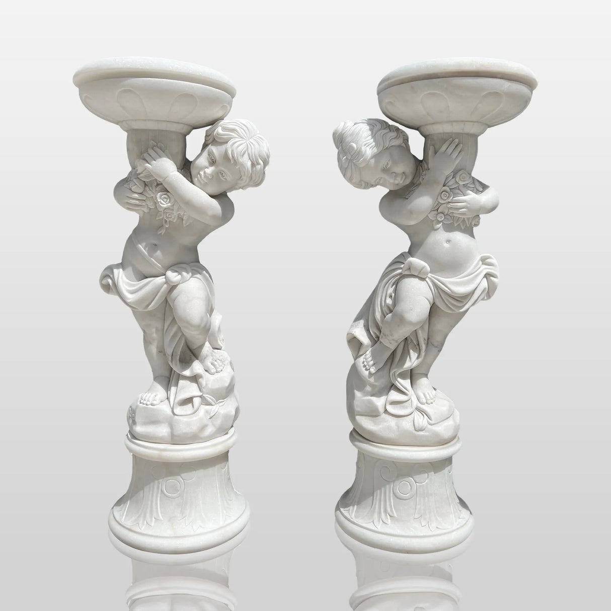 Pair of marble sculptures depicting children holding flowerpots line a curved gravel garden path beside trimmed hedges and rose blooms.