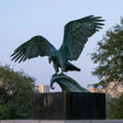 Verdigris bronze eagle statue with broad outstretched wings perches on curved scroll base against outdoor city background.
