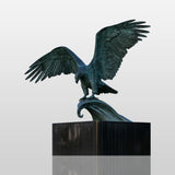 Verdigris bronze eagle statue with broad outstretched wings perches on curved scroll base against outdoor city background.