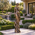 Bronze lady statue with a flower stands on stone patio amid lush landscaped greenery.