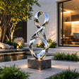 Polished stainless steel ribbon sculpture rests beside a garden water pond.