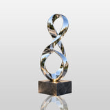 Polished stainless steel ribbon sculpture rests beside a garden water pond.