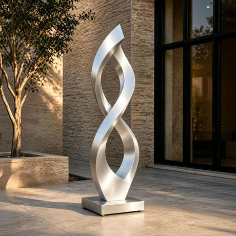 A brushed stainless steel infinity sculpture stands on paved ground outside a modern house.