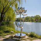 Polished stainless steel lotus sculpture stands on the grassy lake shore under bright sunshine.