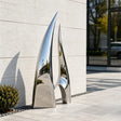 A polished stainless steel sail sculpture sits beside a modern building.