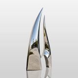 A polished stainless steel sail sculpture sits beside a modern building.