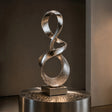 Mirror polished stainless steel ribbon sculpture stands on a square base in dimly lit indoor space.