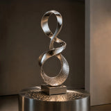 Mirror polished stainless steel ribbon sculpture stands on a square base in dimly lit indoor space.