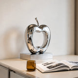 Polished hollow stainless steel apple sculpture stands on a light indoor tabletop.