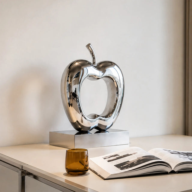 Polished hollow stainless steel apple sculpture stands on a light indoor tabletop.