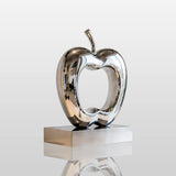 Polished hollow stainless steel apple sculpture stands on a light indoor tabletop.