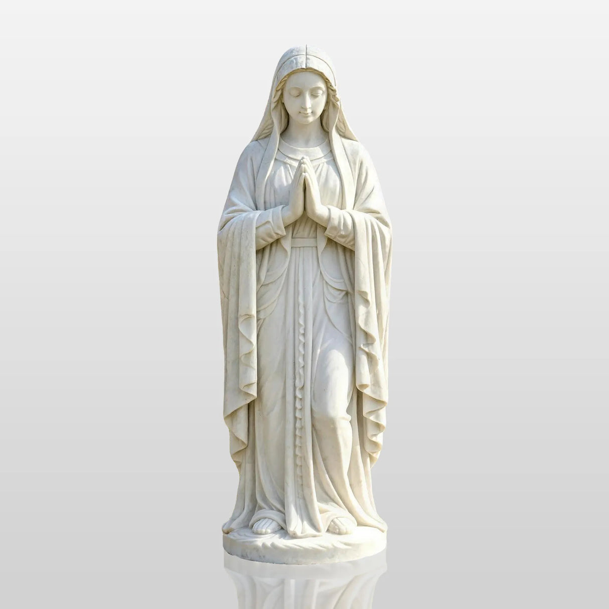White marble Virgin Mary statue stands on paved circle amid green lawn and trees.