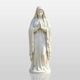 White marble Virgin Mary statue stands on paved circle amid green lawn and trees.