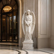 White marble angel statue with wings stands on tiered marble pedestal beside tall stone wall.