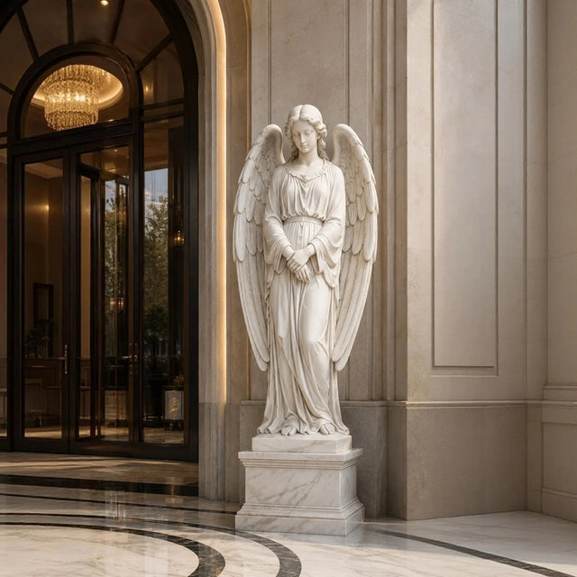 White marble angel statue with wings stands on tiered marble pedestal beside tall stone wall.