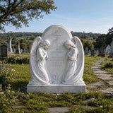 White marble tombstone carved with two kneeling praying angels and central cross sits on green grass within a cemetery.